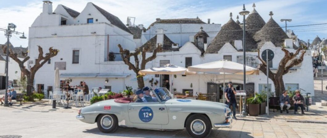 A 1950s open top grey Aston Martin sportscar stops in front of a whitewashed cafe in Bari,