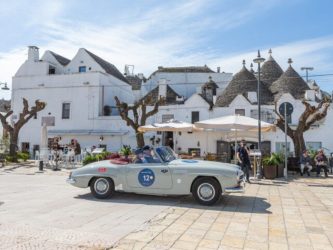 A 1950s open top grey Aston Martin sportscar stops in front of a whitewashed cafe in Bari,