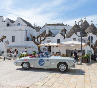 A 1950s open top grey Aston Martin sportscar stops in front of a whitewashed cafe in Bari,