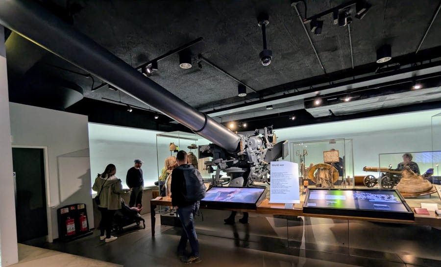 IWM visitors gathered around a large grey gun illuminated in a darkened gallery