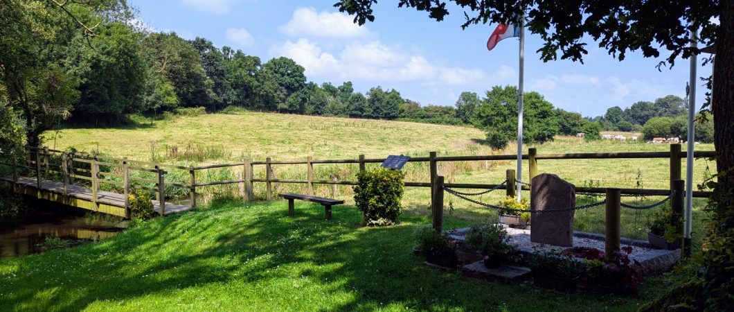 View from a shaded wood across sunny fields with a memorial stone in the foreground