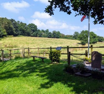 View from a shaded wood across sunny fields with a memorial stone in the foreground