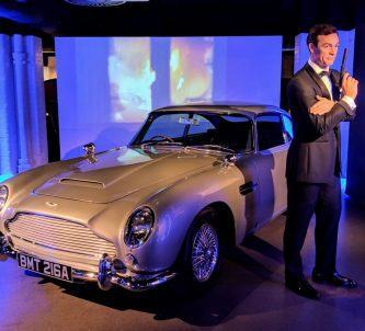 A not-very-lifelike dummy of James Bond in dinner jacket and with a gun, stands next to his silver grey Aston Martin DB5