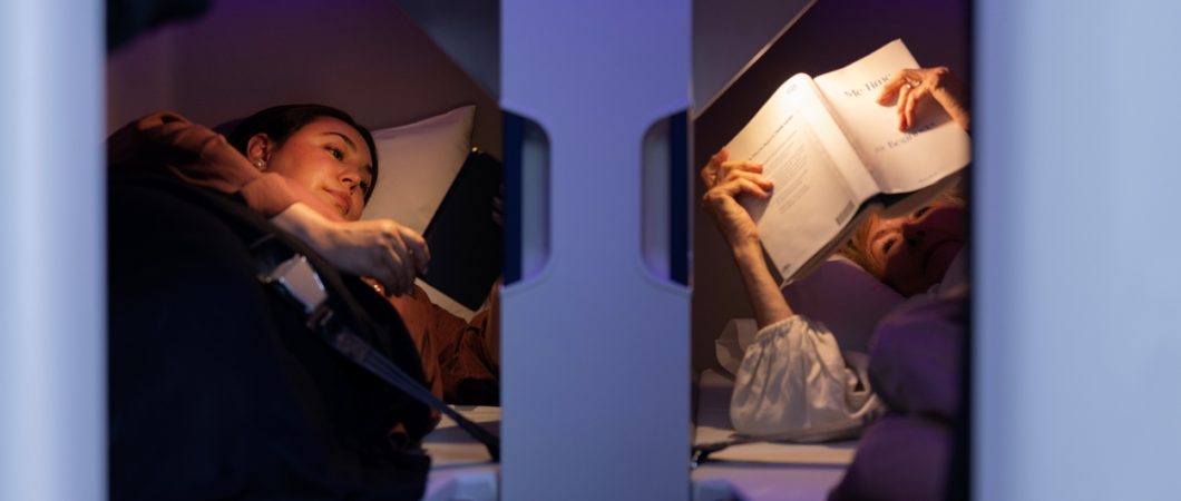 Two passengers lie close to each other in Air New Zealand Skynest bunks under purple lighting