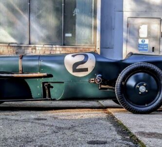 A long green 1920s racing car parked outside a hanger