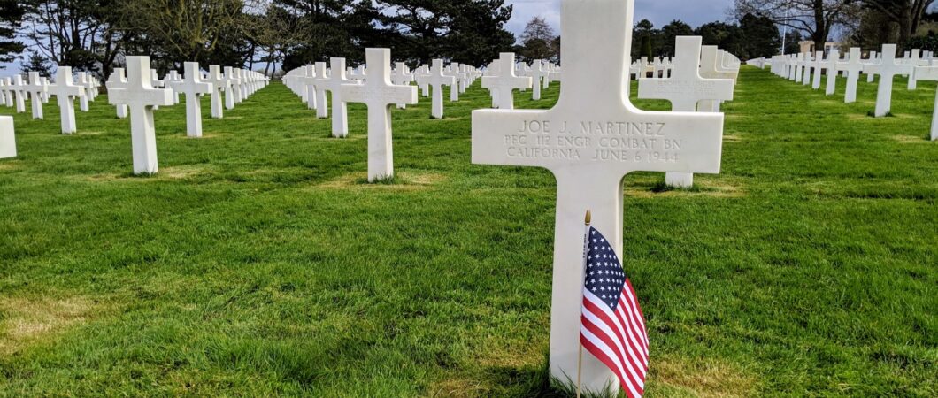 A white cross with a US flag planted in front of it, and many more white crosses stretching away into the distance behind it. The cross is for Joe J. Martinez from California, a private in the 112th combat engineering battalion