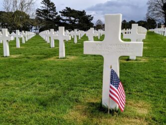 A white cross with a US flag planted in front of it, and many more white crosses stretching away into the distance behind it. The cross is for Joe J. Martinez from California, a private in the 112th combat engineering battalion
