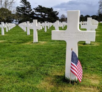 A white cross with a US flag planted in front of it, and many more white crosses stretching away into the distance behind it. The cross is for Joe J. Martinez from California, a private in the 112th combat engineering battalion