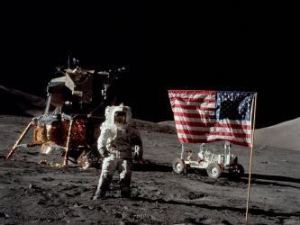 Astronaut stands on the moon in front of their landing capsule, moon buggy, and an American flag