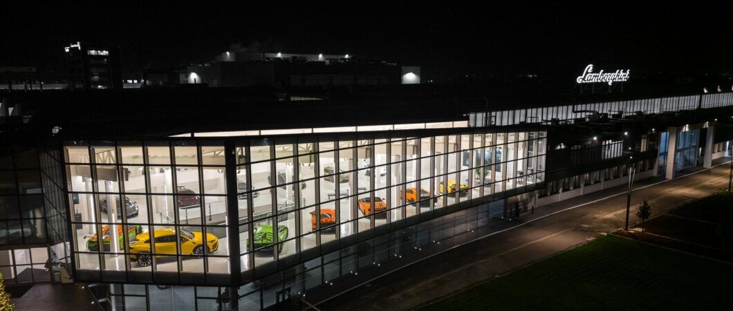 The Lamborghini museum at night. Rows of colourful supercars can be seen in the white-walled setting through the wall-to-ceiling glass windows