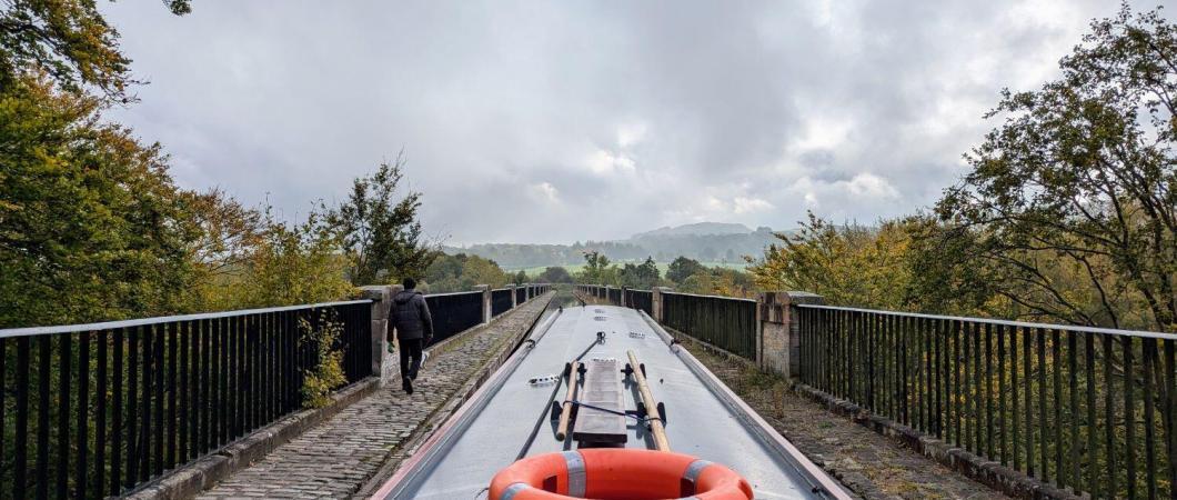 View from the back of a narrowboat along a stone aquaduct above the trees below. A man is walking ahead on the footpath