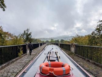 View from the back of a narrowboat along a stone aquaduct above the trees below. A man is walking ahead on the footpath