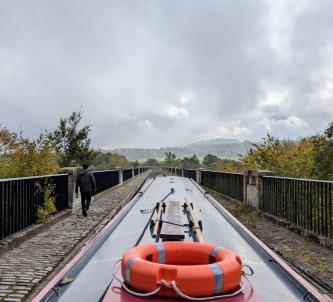 View from the back of a narrowboat along a stone aquaduct above the trees below. A man is walking ahead on the footpath