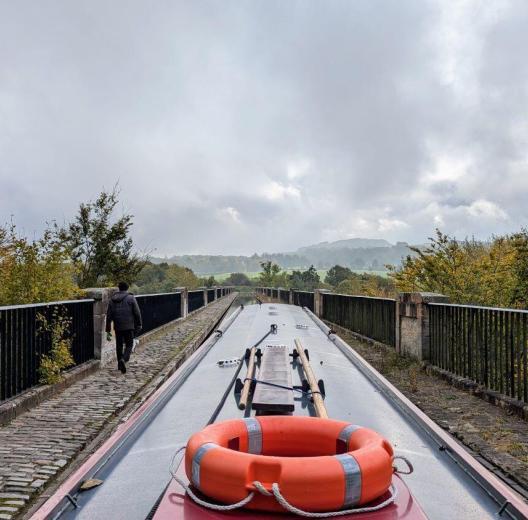 View from the back of a narrowboat along a stone aquaduct above the trees below. A man is walking ahead on the footpath