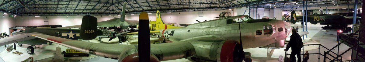 A panorama view of the, fairly dark, hanger with a silver four-engined  B-17 Flying Fortress with a yellow tail and red painted engine nacelles in the foreground