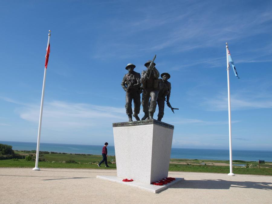 Statue of three soldiers high on a plinth with flagpoles on each side. In the background a man walks past, giving a sense of scale, and there are views, under a clear blue sky, out to sea