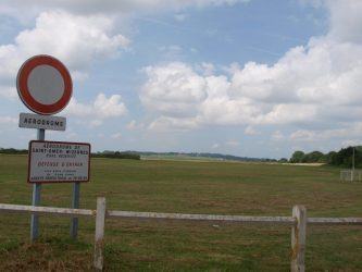 A no entry sign stands in front of a grass airfield with a windsock in the distance