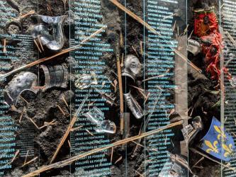 A wall display of broken armour, weapons & flags in mud with an overlay of names in white print
