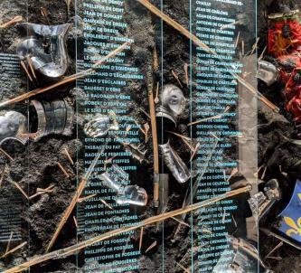 A wall display of broken armour, weapons & flags in mud with an overlay of names in white print