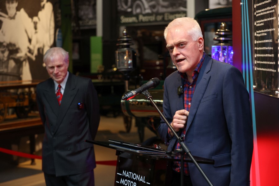 TV presenter Jon Bentley speaking at a microphone with Lord Montagu to his left and classic cars on display behind them.