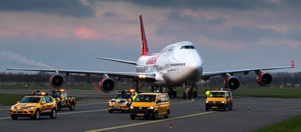 A jumbo jet moving on a taxiway surrounded by yellow vehicles