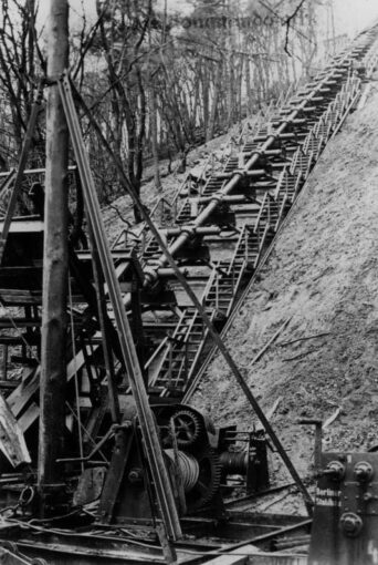 Black and white wartime photo of a long metal barrel with short pipes on both sides at regular intervals, laid up the side of a hill