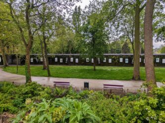 A long low, dark walled, concrete bunker with art posters on the walls at regular intervals, sits among the trees and grass of a municipal park