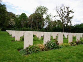 Rows of gravestones at a CWGC cemetery in Normandy. The grassy cemetery is surrounded by large trees.