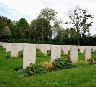 Rows of gravestones at a CWGC cemetery in Normandy. The grassy cemetery is surrounded by large trees.