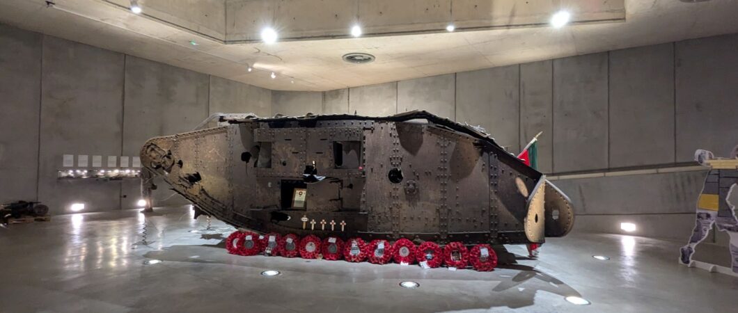 Rusty brown wreckage of a WW1 tank, set in a concrete display hall. A row of red wreaths are laid in front of it.