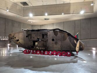 Rusty brown wreckage of a WW1 tank, set in a concrete display hall. A row of red wreaths are laid in front of it.