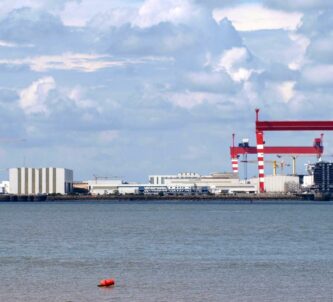 A view across the water of the shipyard, cruise ships under construction, and two giant red gantry cranes