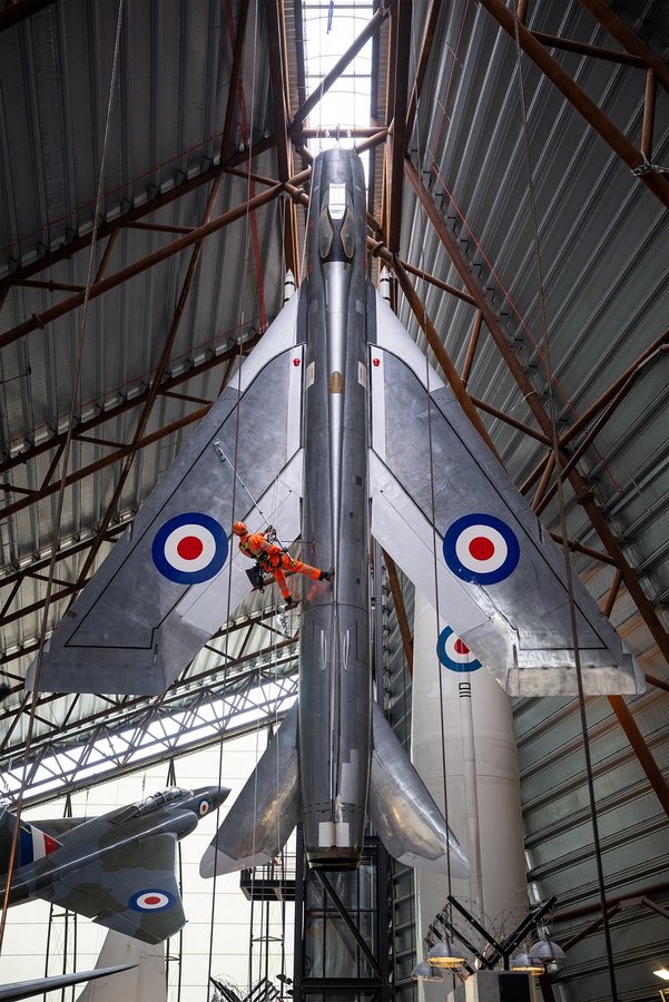 A man in an orange suit suspended on a rope, dusts the wing of a silver English Electric Lightning aircraft that is hanging vertically at the RAF Museum Cosford
