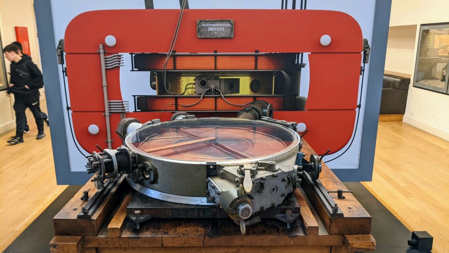 A large circular machine on a table with a red wall and transformer coils behind it