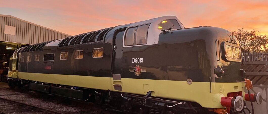 Gleaming diesel locomotive in brown and yellow colours, highlighted by the setting sun, standing in front of a large railway shed