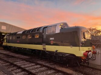 Gleaming diesel locomotive in brown and yellow colours, highlighted by the setting sun, standing in front of a large railway shed