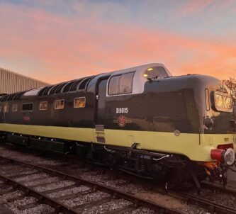 Gleaming diesel locomotive in brown and yellow colours, highlighted by the setting sun, standing in front of a large railway shed