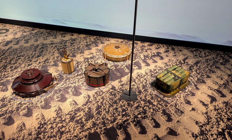 A patch of desert sand with tyre tracks on it and five different type of mines laid out on it.