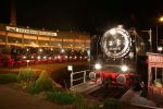Large black steam locomotives are lined up at night in a crescent in front of a roundhouse, with their lights on