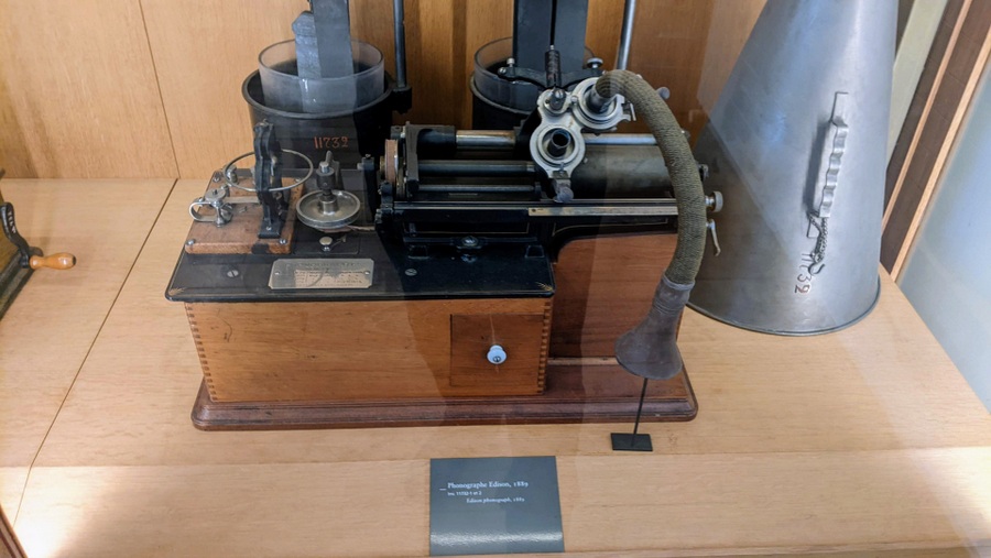 The forerunner of records. A wooden box with, instead of a disc, a black wax tube which revolved and moved so that the needle ran a helix course over it. A speaking tube is attached and a small trumpet/megaphone for playback stands next to it in the display cabinet