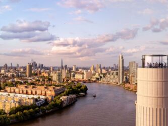 Aerial view of the glass Chimney Lift with people inside at the top of one of Battersea Power Station's chimneys. Behind an below is a panorama of the Thames and London on a glorious sunny day