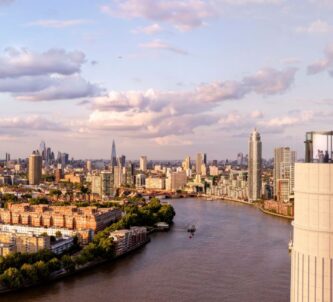Aerial view of the glass Chimney Lift with people inside at the top of one of Battersea Power Station's chimneys. Behind an below is a panorama of the Thames and London on a glorious sunny day