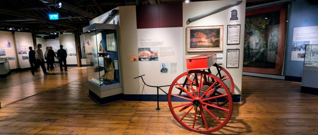 A wooden floored display gallery in the London Museum, Docklands. There are people in the background and a bright red 19th century fire cart in the foreground