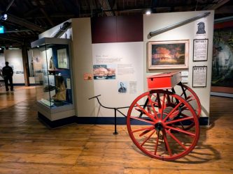 A wooden floored display gallery in the London Museum, Docklands. There are people in the background and a bright red 19th century fire cart in the foreground