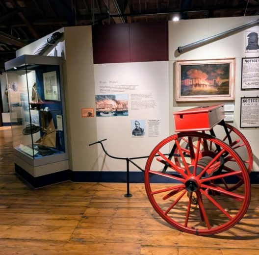 A wooden floored display gallery in the London Museum, Docklands. There are people in the background and a bright red 19th century fire cart in the foreground