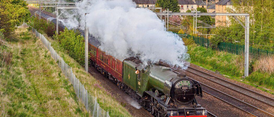 The Flying Scotsman locomotive steams along the tracks with the skyline of Edinburgh behind