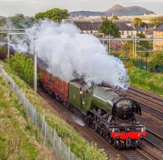 The Flying Scotsman locomotive steams along the tracks with the skyline of Edinburgh behind