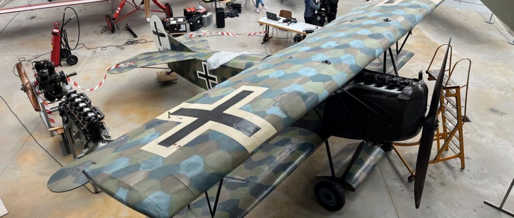 A camouflaged WW1 biplane in a hanger with other aircraft behind