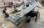 A camouflaged WW1 biplane in a hanger with other aircraft behind