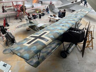 A camouflaged WW1 biplane in a hanger with other aircraft behind